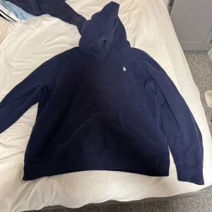 Mörkblå hoodie från Ralph Lauren - Säljer en snygg mörkblå hoodie från Ralph Lauren med klassisk logga på bröstet. Perfekt för en avslappnad stil med känguruficka framtill och bekväm huva. Passar bra till både jeans och joggers.
