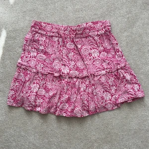 Rosa blommig kortkjol - Charmig rosa kortkjol med ett vackert blommigt mönster. Kjolen har en volangdetalj och elastisk midja för en bekväm passform. Perfekt för en somrig look! Obs! De är shorts på insidan av kjolen (passar nog både XS,S)