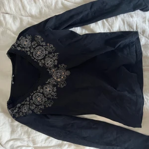 Svart långärmad topp med broderi - Säljer en svart långärmad topp med v-ringning och detaljerat vitt broderi framtill. Perfekt för dig som vill ha något unikt och snyggt i garderoben. Passar till många olika stilar!
