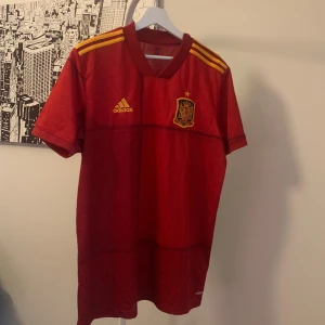 Spansk landslagströja från Adidas - Röd fotbollströja från Adidas med Spaniens landslagsmärke på bröstet och gula detaljer på axlarna. Klassisk V-ringning och korta ärmar. Perfekt för dig som vill visa ditt stöd för Spanien på matchdagen.