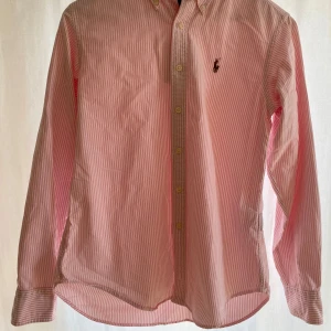 Rosa och vit randig skjorta från Ralph Lauren - Snygg rosa och vit randig skjorta från Ralph Lauren med klassisk passform. Skjortan har långa ärmar och knappar framtill samt en broderad logotyp på bröstet. Perfekt för en stilren look. Storlek S passar XS (pris ej hugget i sten🪨💸)
