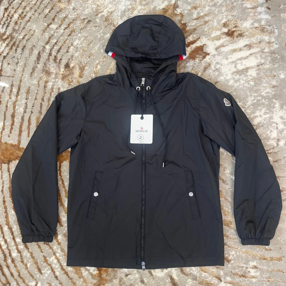 Snygg svart vindjacka från Moncler med huva och dragkedja. Jackan har elastiska ärmslut och två fickor med knappar framtill. Perfekt för blåsiga dagar.. Takit.