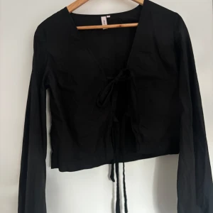 Svart knytblus med långa ärmar - Säljer en svart blus med långa ärmar och knyt framtill. Blusen har en djup v-ringning och är croppad i modellen. Perfekt att styla till olika outfits.