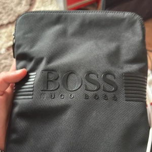 Svart axelväska från Hugo Boss - Snygg svart axelväska från Hugo Boss med tydlig logga framtill. Väskan har dragkedja upptill och är perfekt för att bära med sig det viktigaste. Stilren design som passar till många tillfällen.