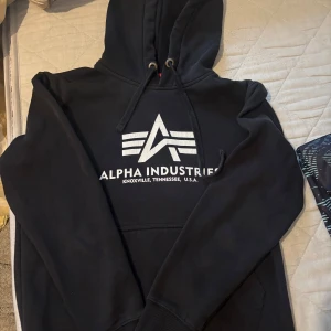Svart hoodie från Alpha Industries - Säljer en svart hoodie från Alpha Industries med vit logga och text på bröstet. Tröjan har huva och en stor magficka framtill. 