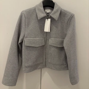 Grå jacka med dragkedja från Kappahl - Snygg grå jacka från Kappahl med dragkedja framtill och två stora fickor. Jackan har klassisk krage och raka, långa ärmar. Perfekt för dig som gillar stilren och enkel design. Helt ny aldrig använd med prislapp.