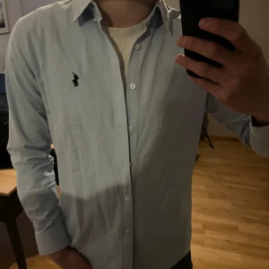 Ljusblå skjorta från Ralph Lauren - Säljer en klassisk ljusblå skjorta från Ralph Lauren med broderad logga på bröstet. Skjortan har lång ärm, knappar framtill och en stilren krage. Perfekt för dig som gillar en tidlös och clean look.