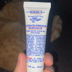 Kiehl's Ultimate Strength Hand Salve - En tub med Kiehl's Ultimate Strength Hand Salve, en intensivt återfuktande handkräm för mycket torra och aktiva händer. Innehåller 30 ml och är perfekt att ha med sig i väskan. Krämen är vit och kommer i en smidig plasttub med blå och röd text.