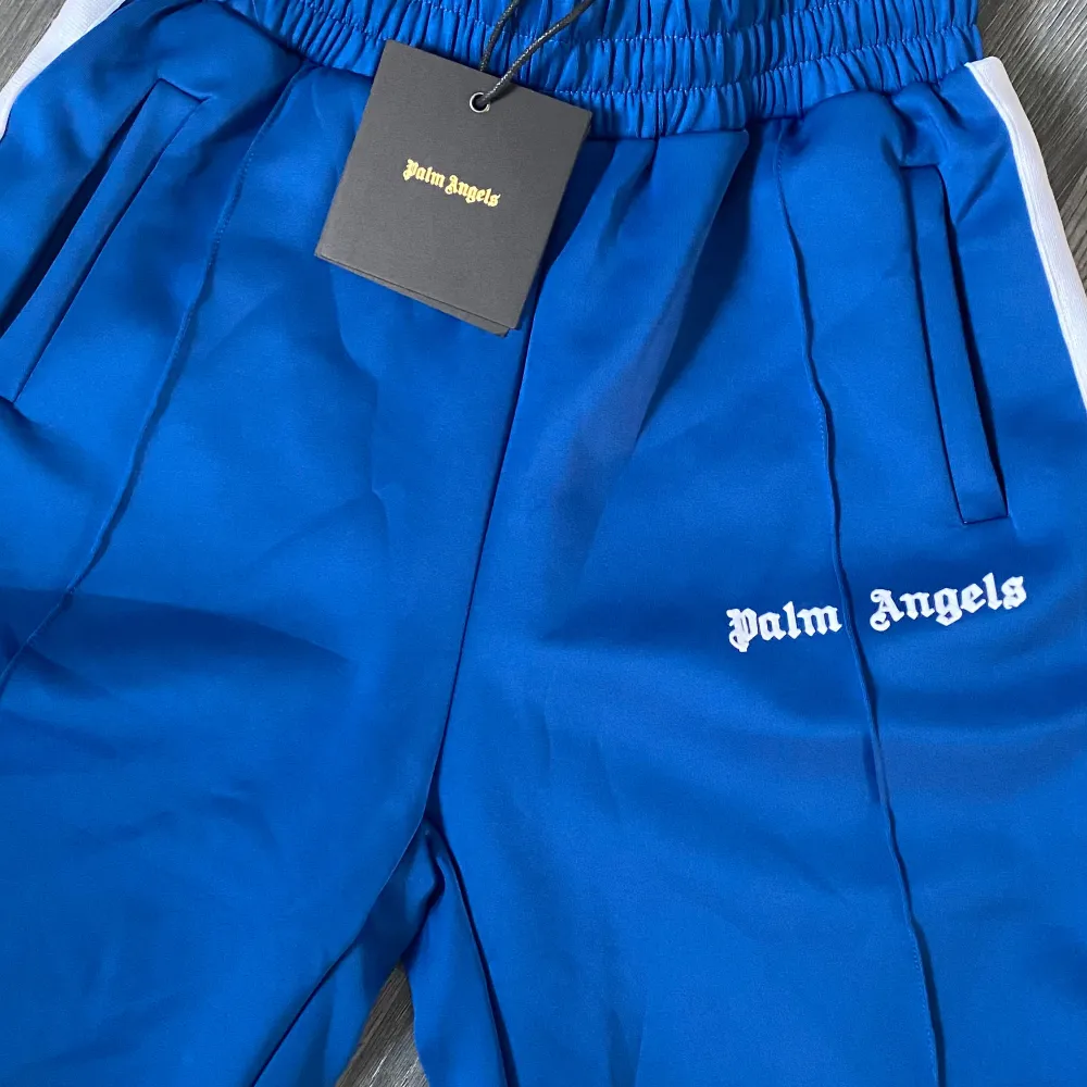 Snygga blå träningsbyxor från Palm Angels med vita sidor och logga på ena benet. Byxorna har resår i midjan och raka ben med fickor på sidorna. Perfekta för en sportig och trendig look.. Shortsit.