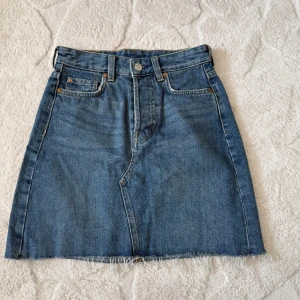 Jeanskjol med rå kant - Säljer en klassisk blå jeanskjol med rå, fransig kant nertill. Kjolen har fem fickor, bälteshällor och knappstängning framtill. Perfekt basplagg för garderoben och enkel att styla till olika looks.