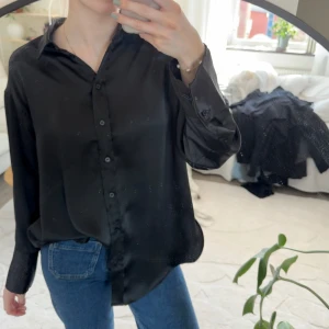 Svart satinblus med knappar - Snygg svart skjorta i glansigt satinmaterial med klassisk krage och knäppning framtill. Långa ärmar med manschett och avslappnad passform. 