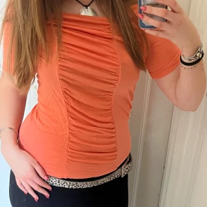 Orange topp med rynkdetaljer - Säljer en snygg orange topp med rynkdetaljer framtill. Toppen har korta ärmar och en figurnära passform. Toppen har en liten fläck som syns på sista bilden. Skriv vid frågor!🤗😍