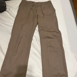 Beiga chinos med dragsko - Säljer ett par beiga chinos med dragsko i midjan och raka ben. Byxorna har dragkedja och knapp framtill samt två bakfickor. Perfekta för en avslappnad men ändå stilren look. Ny pris på dessa var runt 2000. Om ni har fler frågor så är det bara att skriva:) 