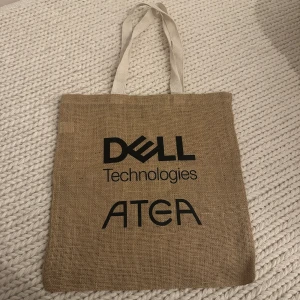Beige tygkasse med Dell Technologies och Atea-logga - Säljer en beige tygkasse i grovt tyg med svarta tryckta loggor från Dell Technologies och Atea. Väskan har långa vita handtag och är perfekt för att bära böcker, mat eller annat smått och gott.