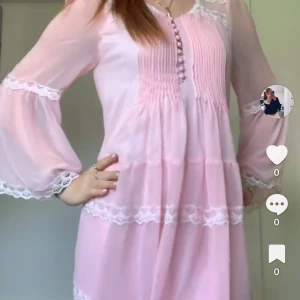 Ljusrosa klänning med spetsdetaljer från Club L - Romantisk ljusrosa klänning med långa, vida ärmar och spetsdetaljer. Klänningen har knappar framtill, markerad midja med knytband och volangkant nertill. Perfekt för vår och sommar. Första bilden visar färgen bäst. Endast använd en gång. 