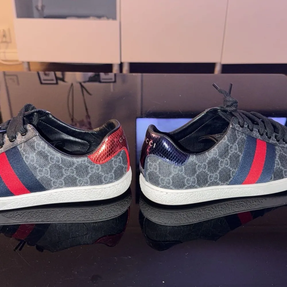 Snygga Gucci sneakers med det klassiska monogrammönstret i grått och svarta snören. Skorna har röda och blå ränder på sidorna och metallic-detaljer i rött och blått på hälen. Perfekta för en stilren look.. Kengät.