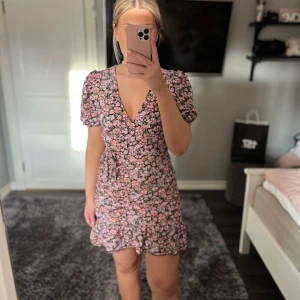 Blommig kortklänning med volang - Söt blommig kortklänning i rosa och svart med v-ringning och volangdetaljer. Klänningen har korta ärmar och en figurnära passform som ger en feminin siluett💗