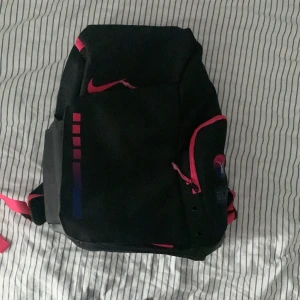 Svart och rosa ryggsäck från Nike - Snygg svart ryggsäck från Nike med rosa detaljer och justerbara axelremmar. Väskan har flera fack med dragkedja och en cool logga på framsidan. Perfekt för skolan eller träningen!