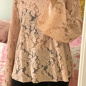 Rosa spetsblus med vida ärmar från Lindex - Säljer en ljusrosa blus i spets från Lindex med blommigt mönster och vida långa ärmar. Blusen har en rund halsringning med dekorativa band i nacken och är genombruten i materialet. Perfekt för dig som gillar romantisk stil!