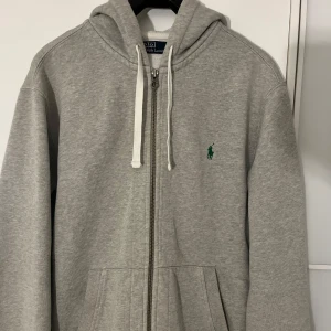 Grå zip från Polo Ralph Lauren - Säljer en klassisk grå hoodie från Polo Ralph Lauren med dragkedja och vit dragsko. Tröjan har en broderad grön logga på bröstet och två fickor framtill. Perfekt för en avslappnad stil.
