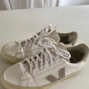 Vita sneakers från Veja med grå detaljer 39 - Säljer ett par klassiska vita sneakers från Veja med grå V-logga på sidan och grå detaljer vid hälen. Skorna har snörning och platt sula, perfekt för en stilren vardagslook. Bra skick utvändigt, trasiga invändigt i hälen (mer på höger), går att fixa med hälskydd fr skoaffär som klistras fast, kostar ca 30-50kr/par.