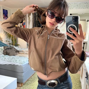 Brun jacka med huva från Odd Molly - Söt ljusbrun jacka från Odd Molly med huva, justerbart band i midjan och dragkedja. Jackan har vita kontrastsömmar, två fickor fram och ett litet hjärtmärke på ärmen. Perfekt nu på våren!🌸