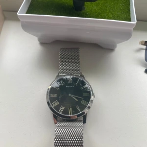 Silverfärgad klocka från Guess med mesh-armband - Stilren klocka från Guess med svart urtavla, romerska siffror och ett elegant silverfärgat mesh-armband i metall. Klassisk design som passar till många tillfällen. Urtavlan är rund och boetten är också i silver. Den har en liten repa men går att fixa!