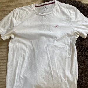 Vit t-shirt från Hollister - Klassisk vit t-shirt från Hollister med liten broderad logga på bröstet. T-shirten har rund halsringning med vinröd detalj på insidan och är tillverkad i mjuk bomull. Perfekt för en enkel och stilren look.