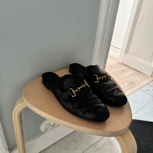 Svarta loafers med gulddetaljer - Snygga svarta slip-in loafers i äkta skinn med guldfärgade detaljer på ovansidan. Perfekta för en stilren look. Originalpris 1500kr.