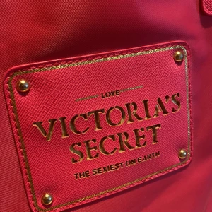 Rosa väska från Victoria's Secret - Säljer en snygg rosa shoppingväska från Victoria's Secret med stort guldigt logomärke framtill. Väskan har två rejäla handtag och rymligt svart innerfoder. Perfekt för att bära med sig allt man behöver under dagen.