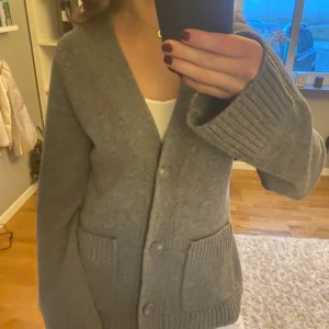 Grå kofta från Wera - Säljer en stilren grå kofta från Wera. Koftan har en v-ringad design med knappar framtill och två praktiska fickor. Den är långärmad med ribbade muddar vid ärmslut och nederkant, vilket ger en klassisk look. Nypris: 899 kr