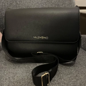 Svart axelväska från Valentino - Snygg svart axelväska från Valentino i elegant design. T:er på väskan har ramlat av, jag tror säkert att man kan besöka valentino butiken och få det ersatt. Sedan kan man även beställa hem ett nytt T som går att ersätta det gamla. Skriv gärna vid intresse eller frågor!🩷🌸