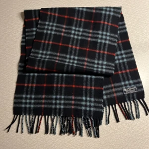 Burberry halsduk - Snygg rutig halsduk från Burberry i svart, grått och rött. Tillverkad i mjukt material med klassiskt rutmönster och fransar i ändarna. Perfekt för att hålla stilen i kyligare väder.