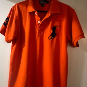 Orange pikétröja från Ralph Lauren - Säljer en snygg orange pikétröja från Ralph Lauren med korta ärmar och klassisk krage. Tröjan har en broderad logga på bröstet och siffran 3 på ärmen. Perfekt för en sportig look!