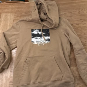 Beige hoodie från H&M - Säljer en beige hoodie från H&M med en tryckt bild på framsidan. Den har en klassisk känguruficka och justerbar huva med snören. Perfekt för en avslappnad stil. Använd 1 gång
