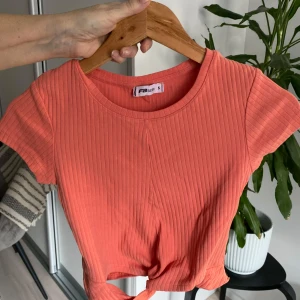 Korallfärgad ribbad topp  - Säljer en snygg korallfärgad ribbad topp Toppen har korta ärmar och en knytdetalj framtill som ger en trendig look. Perfekt för varmare dagar!