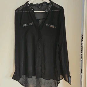 Svart blus med nitar - Snygg svart blus med nitar på kragen och bröstfickor. Blusen har långa ärmar och knappar framtill. Perfekt för en edgy look!