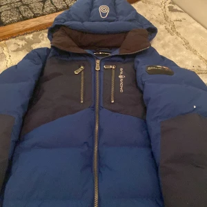 Blå dunjacka från Sail Racing - Säljer en snygg blå dunjacka från Sail Racing med dragkedja och två bröstfickor. Jackan har en cool patch på ärmen och är perfekt för kalla dagar. Den har en hög krage och justerbar huva för extra skydd mot väder och vind.