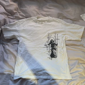 Vit t-shirt med tryck från Abundant brands - Säljer en vit t-shirt med ett coolt tryck fram och bak. Framsidan har en mystisk figur med kors och baksidan visar ett detaljerat slott med text ovanför. Perfekt för dig som gillar unika och konstnärliga plagg!