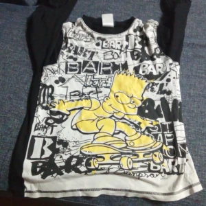 Svart och vit långärmad tröja med Bart Simpson - Cool långärmad tröja med svart bas och vit framsida prydd med ett stort tryck av Bart Simpson på skateboard. Tröjan har ett grafiskt mönster med text och illustrationer i svart och gult. Storleken 146-152 11-12 år