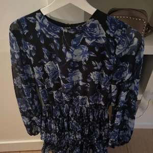 Blommig klänning med långa ärmar - Snygg klänning med blått blommönster på svart bakgrund. Den har långa ärmar och en smickrande passform med elastisk midja. Perfekt för en stilfull look.