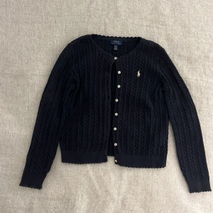 Mörkblå kofta från Ralph Lauren - Säljer en jättefin mörkblå kofta från Ralph Lauren med kabelstickat mönster och knappar framtill. Ca 12-14år❣️100% bomull! Den har en klassisk design som passar perfekt till olika tillfällen. Inga fläckar finns❣️❣️ på lagningar på armen men de syns inte!!