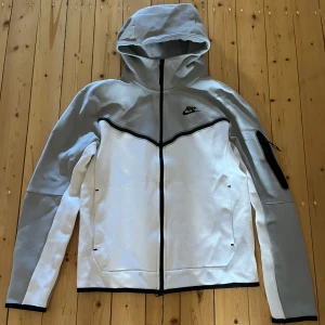 Nike Tech Fleece grå och vit  - Säljer min grå vita Nike tech fleece som inte används mycket. Lite nopprig fram vid ärmen och har en liten fläck som inte går bort vid ena ärmen. Men annars fin kvalité! 