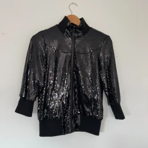 Svart glittrig jacka med paljetter - Snygg svart jacka täckt med glittriga paljetter. Jackan har en dragkedja framtill och trekvartsärmar. Perfekt för att sticka ut på festen!
