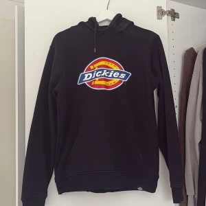 Svart hoodie från Dickies - Säljer en svart hoodie från Dickies med deras ikoniska logga i färgglatt tryck på framsidan. Tröjan har en klassisk passform med långa ärmar och en praktisk magficka. Perfekt för en avslappnad stil.
