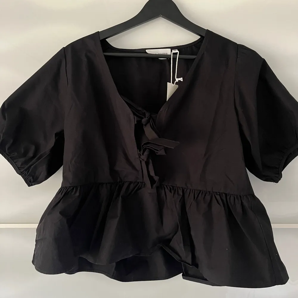 Snygg svart blus från Carin Wester med korta puffärmar och v-ringning. Blusen har knytband framtill och en volangdetalj vid nederkanten. Perfekt för en stilren look.. Yläosat.