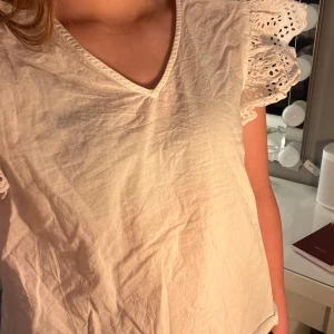 Vit blus med korta spetsdetaljer - Säljer en söt vit blus med spetsdetaljer på ärmarna. Blusen har en v-ringad hals och korta ärmar med volang. Perfekt till sommaren! 