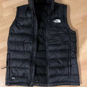 Svart dunväst från The North Face - Säljer en svart dunväst från The North Face med dragkedja och hög krage. Västen har en quiltad design och en bröstficka med dragkedja. Perfekt för våren, västen är använd fåtal gånger. Passar som S/M