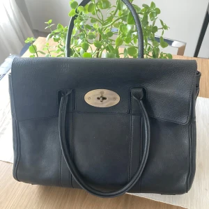 Svart Mulberry handväska med metallspänne, äkta skinn - Elegant svart handväska i skinn med ett stilrent metallspänne framtill. Väskan har två handtag och en praktisk dragkedja inuti för säker förvaring. Perfekt för att bära dina dagliga nödvändigheter med stil.
