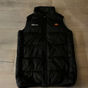 Svart dunväst från Ellesse - Snygg svart dunväst från Ellesse med dragkedja framtill. Perfekt för lager-på-lager under kyligare dagar. Västen har en stilren design med Ellesse-loggan på bröstet.